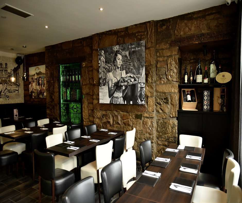 dining room of Mamma Roma Ristorante with stone walls and dark wood tables and chairs. There is a painting of a woman holding a basket of fruit on the wall.