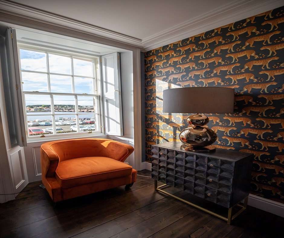 a living room with a large window, an orange armchair, and a tallboy dresser with a silver lamp on it. The walls are covered in a dark blue wallpaper with a pattern of gold leopards.