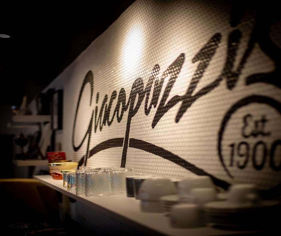 A section of an ice cream shop There is a wall with a black and white logo that says 'Giacopazzi Est. 1900'. There are glass and ceramic cups on a shelf in front of it.