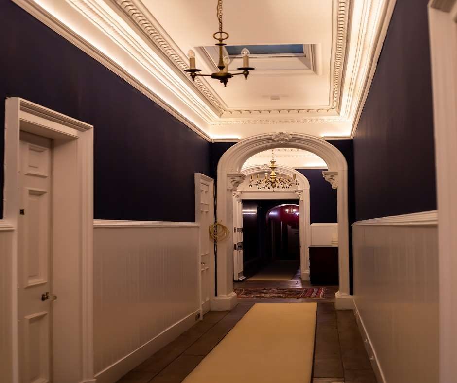 This is a photo of a hallway of the Ayton Castle. The walls are painted dark blue and the floor is covered with a beige carpet. There are two doors on the left side of the hall and one door on the right side. The ceiling is decorated with ornate white trim.