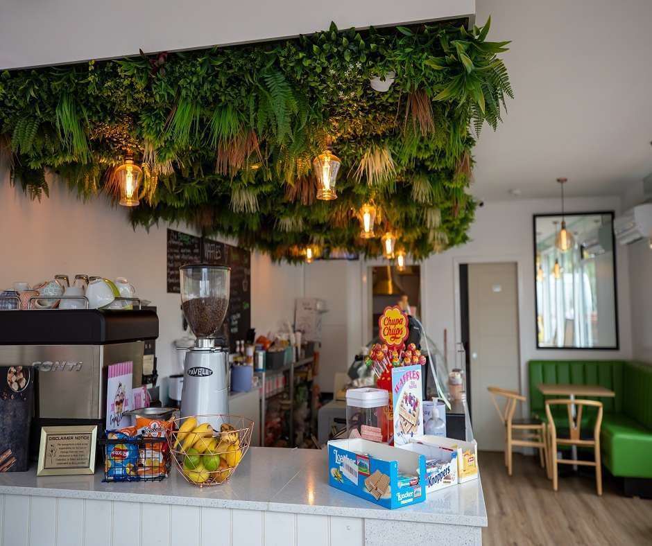 the interior of a cafe, Crêpes and Creams. There is a counter with a coffee machine and some food on it. There are tables and chairs in the background. The ceiling is decorated with fake plants and there are some lights hanging down.