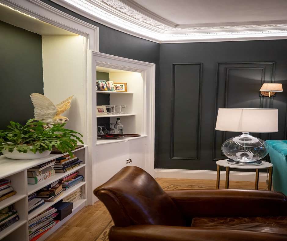 A dark green paneled living room with white trim. There is a brown leather chair, a white lamp, and a globe on a table. There is a bookshelf with a bird statue and plants on it. There is a built-in cabinet with a bar.