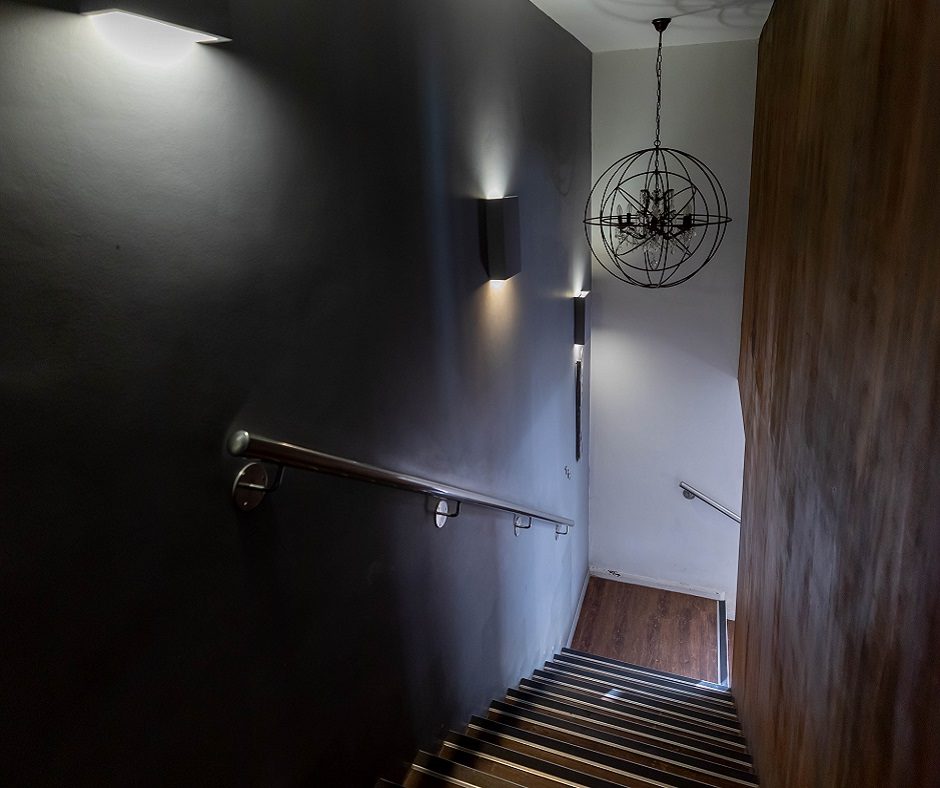 staircase with dark walls and wood steps. There is a metal railing on the left side of the stairs, and two lights on the wall. There is a round light fixture hanging from the ceiling.