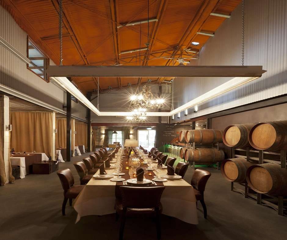 interior of a winery. known as The Barrel Room. There is a long wooden table set with white tablecloths and brown leather chairs. There are wine barrels stacked in the background and a large light fixture hanging from the ceiling.