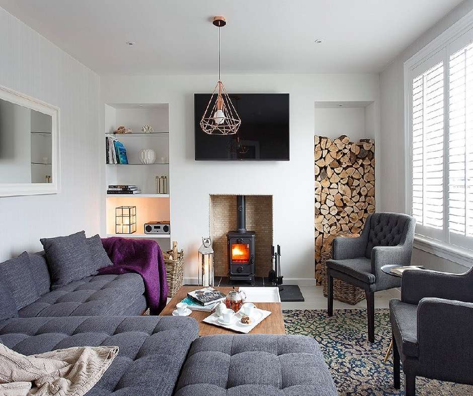 A living room with a gray couch, two gray armchairs, a fireplace, a television, and a woodpile.