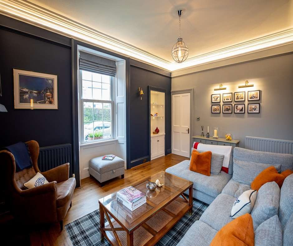 Interior design showing a living room with blue walls, a gray couch, and a brown leather chair. There is a coffee table in front of the couch with books on it, and a gray rug under the coffee table. There is a window on the left side of the couch and a door on the right side of the couch. There are pictures on the wall behind the couch and a lamp on the wall behind the chair.