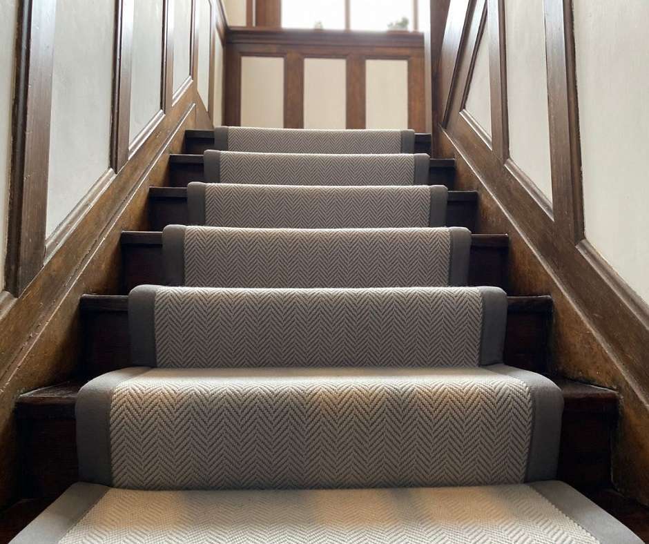 Interior design of a carpeted staircase with wood handrails. The carpet is a light gray herringbone pattern with a solid gray border.