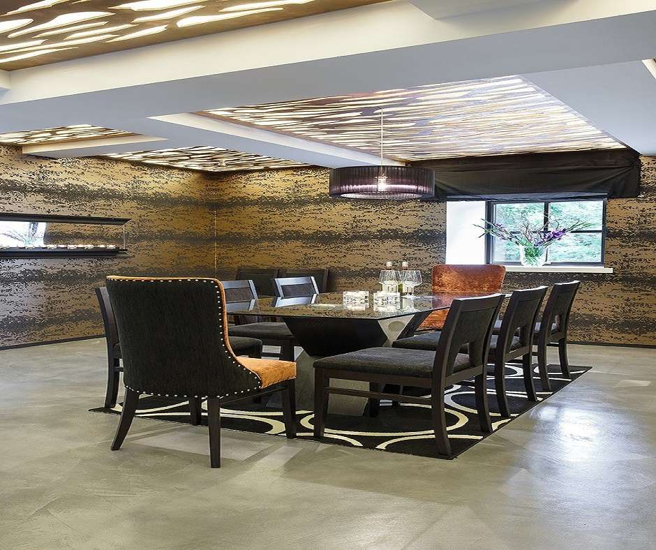 There is a dining room with a large table and several chairs. The walls are decorated with a brown and black wave pattern and the floor is grey with a black and white circular rug in the center. There is a black lampshade hanging from the ceiling which is decorated with several circular cutouts.