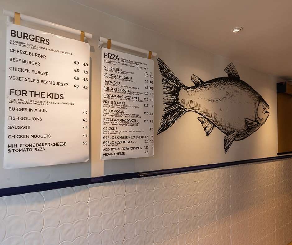 There is a wall with 2 signs on it. One sign lists burgers and their prices. The other lists pizzas and their prices. There is a drawing of a fish on the wall.