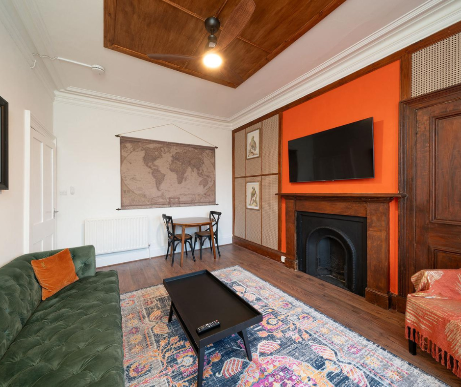 an image of a living room. There is a fireplace, a television, a sofa, and a table with two chairs. The walls are painted white and orange. There is a wooden ceiling with a fan.