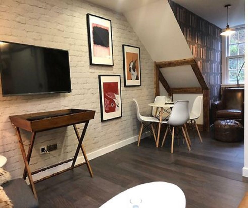 There is a holiday let living room with a TV, a table with four chairs, and a brown leather chair. There are brick patterned wallpaper and several paintings on the walls.
