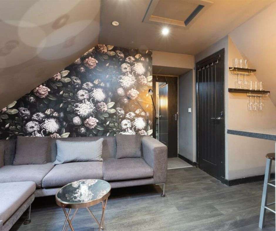 A holiday let living room. There is a floral wallpapered wall behind a gray couch with a copper table in front of it. There is a dark colored door and a partial wall with wine glasses hanging on it to the right of the couch.
