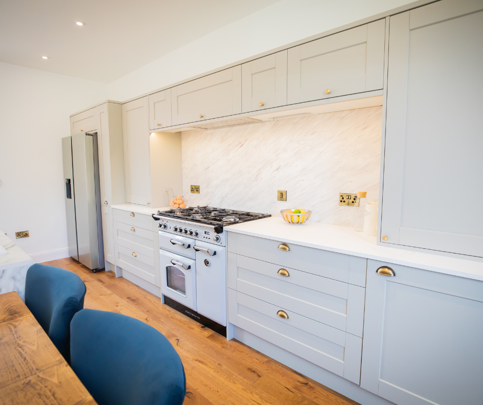 A kitchen with gray cabinets, a large white marble countertop, and a stainless steel oven and refrigerator. The floor is hardwood, and there are two blue chairs at a table in the foreground.