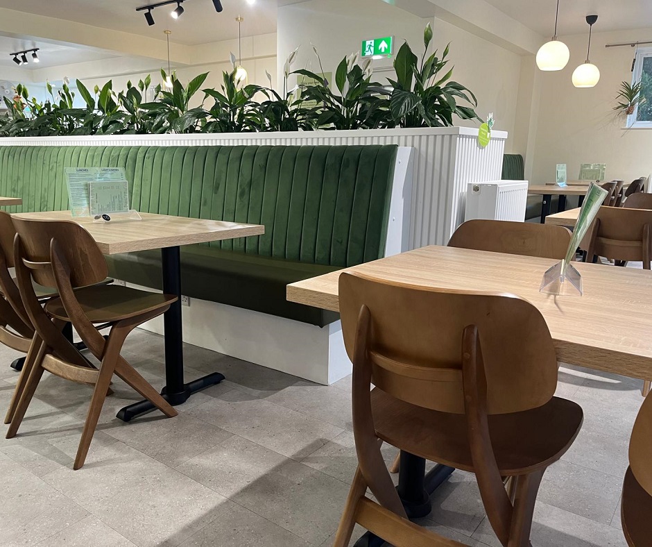 A modern café interior with wooden tables and chairs, green cushioned booth seating, and potted plants on a divider. Warm lighting and neutral colors create a cozy, inviting atmosphere.