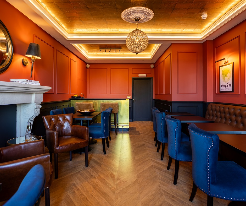Stylish lounge with blue velvet chairs, brown leather armchairs, wooden tables, warm red walls, ornate ceiling lights, and a golden bar at the back. The room features elegant decor and wooden herringbone flooring.