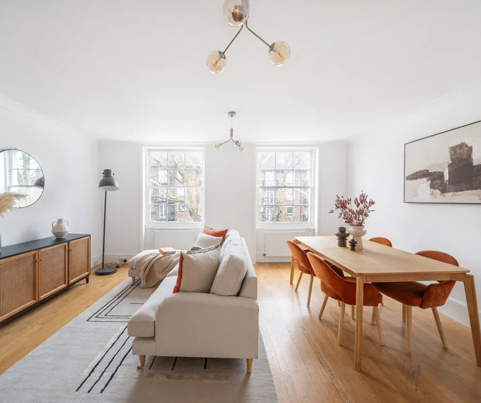 A bright, open-plan living and dining room showcasing a modern aesthetic. It features a light cream sectional sofa and a wooden dining table with vibrant orange chairs.
