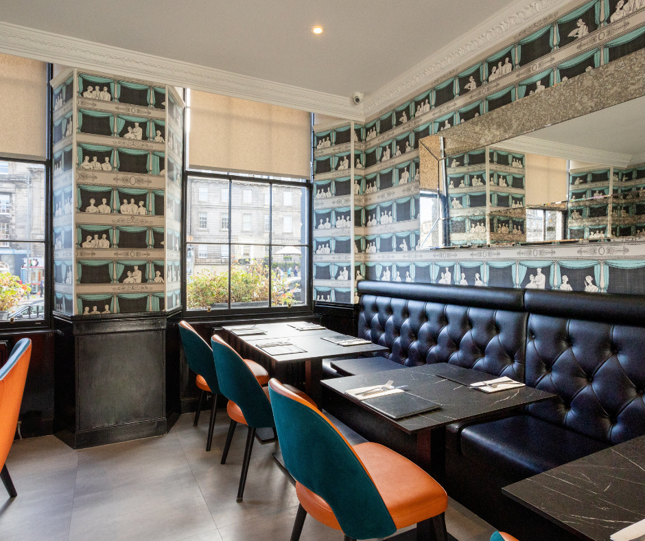 A modern restaurant interior features black booth seating, dark tables, and colorful orange and teal chairs. Patterned wallpaper lines the walls.
