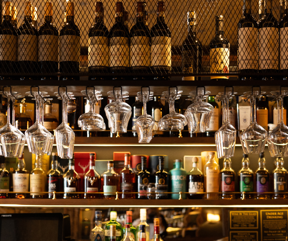 A warm, inviting bar area features shelves stocked with various bottles and two rows of hanging glasses. It highlights a diverse selection of beverages.